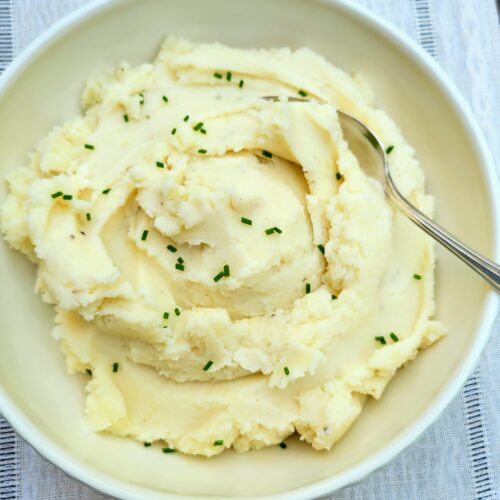 Mashed potatoes with chives in white bowl with spoon on table cloth